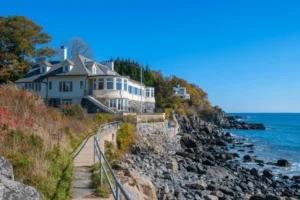 Coastal home perched on a rocky shoreline with a winding seaside path, autumn foliage, and clear blue ocean under a bright sky.