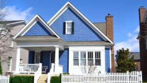 Blue craftsman-style house with white trim, covered front porch, picket fence, and gabled roof under a clear blue sky in a suburban neighborhood.